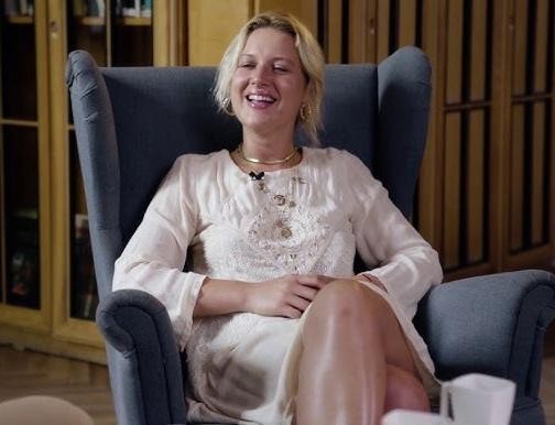 Aleksandra Domańska śmiejąca się na planie filmowym, w tle zamazane rekwizyty i sprzęt filmowy.