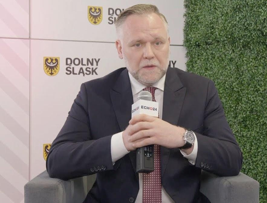 Dawid Jackiewicz przemawia publicznie, gestykulując, w nowoczesnym wnętrzu, symbolizującym jego aktywność polityczną.