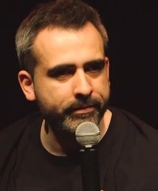 Abelard Giza na scenie podczas występu stand-upowego, trzymający mikrofon, z inteligentnym wyrazem twarzy, w tle rozmyte światła sceny.