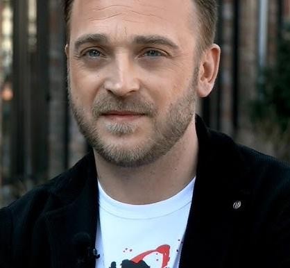 Mateusz Gessler w dynamicznej pozie, gotujący lub wydający polecenia w ruchliwej, nowoczesnej kuchni restauracyjnej, otoczony sprzętem kuchennym i innymi kucharzami.