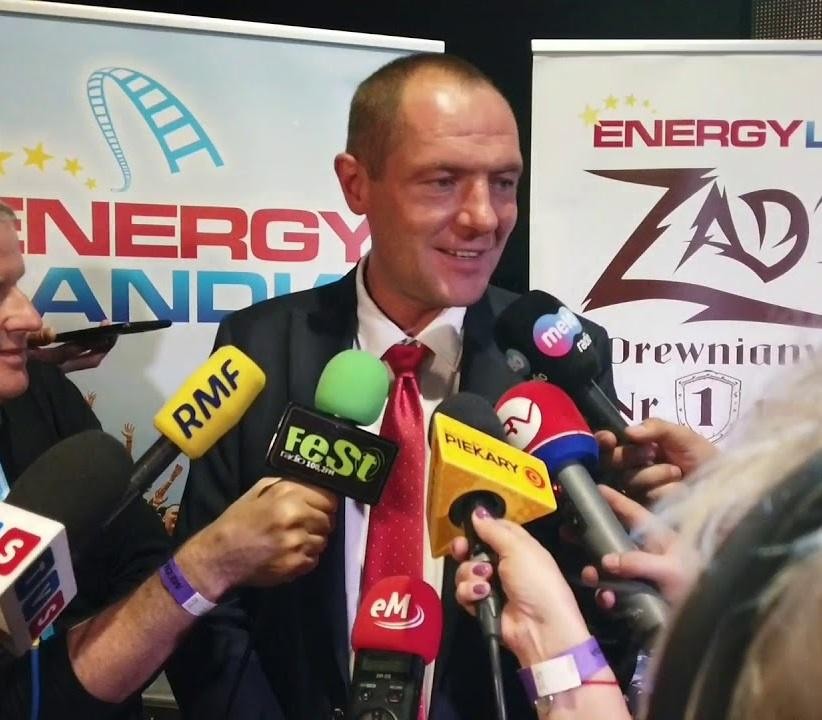 Marek Goczał w stroju rajdowym obok swojego samochodu SSV na tle toru rajdowego.