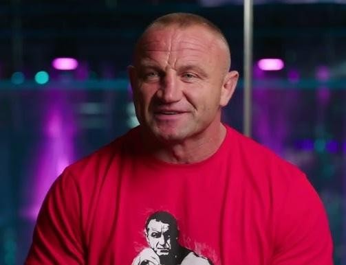 Mariusz Pudzianowski podczas walki MMA w oktagonie, wyprowadzający cios lub w zwarciu, ukazujący intensywność i atletyczną formę.