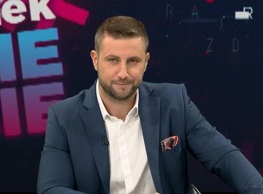 Dynamiczny widok na studio telewizyjne z biurkiem prezentera i ekranem z grafiką informacyjną, symbolizujący środowisko pracy Miłosza Kłeczka.