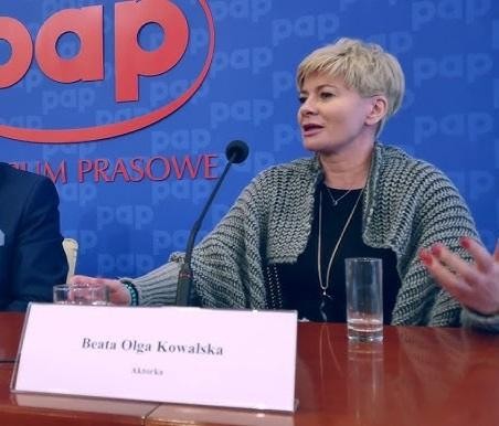 Beata Olga Kowalska w dynamicznej pozie na scenie, oddającej jej wszechstronność jako aktorki i choreografki, z wyrazistą mimiką i gestem, symbolizująca jej bogatą karierę teatralną i filmową.
