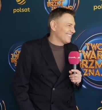 Krzysztof Ibisz w akcji na planie telewizyjnym, dynamicznie prowadzący program, otoczony sprzętem studyjnym, symbolizujący jego rolę jako doświadczonego prezentera.